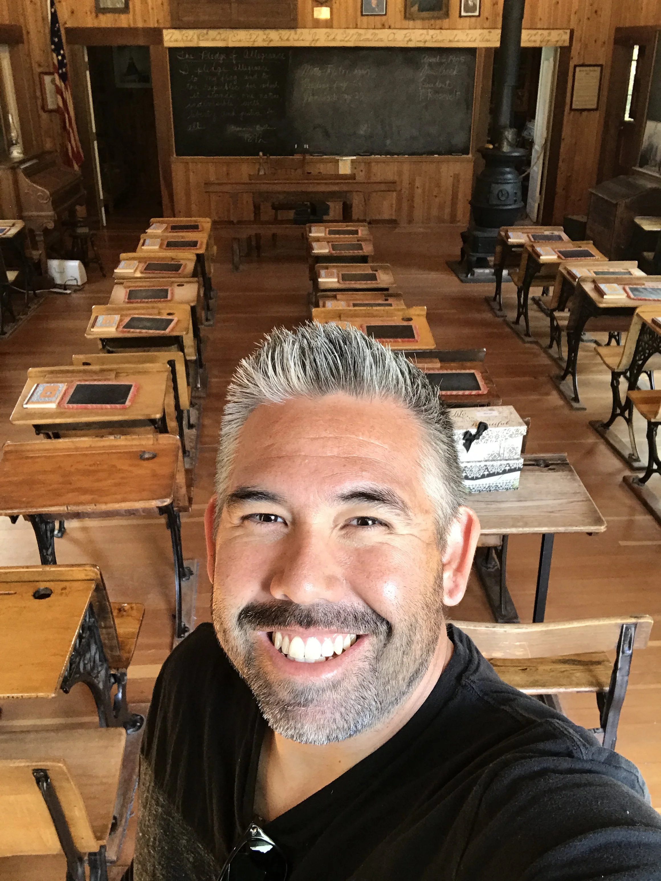 A man with short gray hair and a beard taking a selfie in a wooden classroom with desks, chalkboard, and a wood stove in the background.