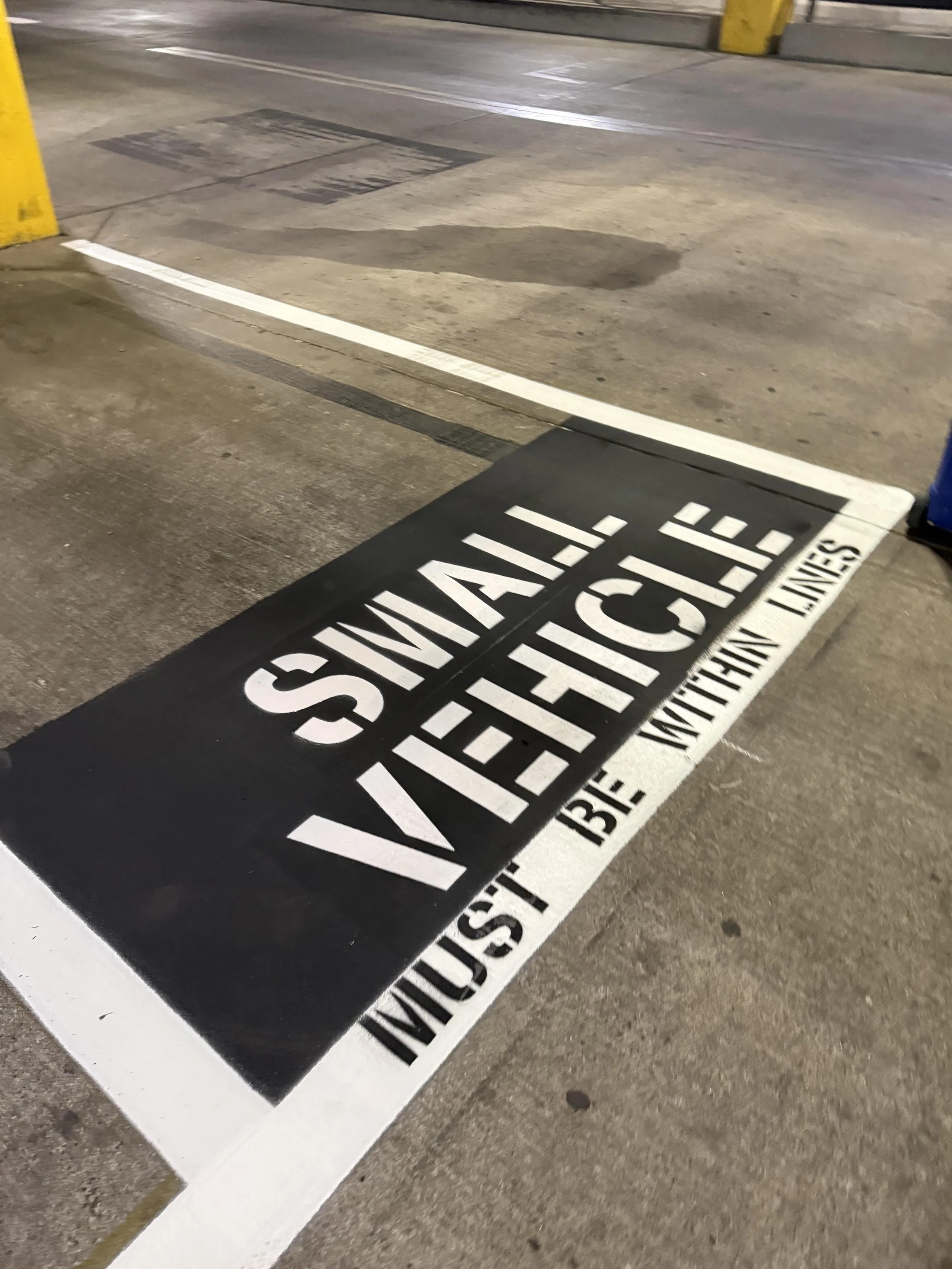 A black and white warning sign on the ground in a parking lot instructing to keep the area clean, with white and yellow parking barriers visible in the background.