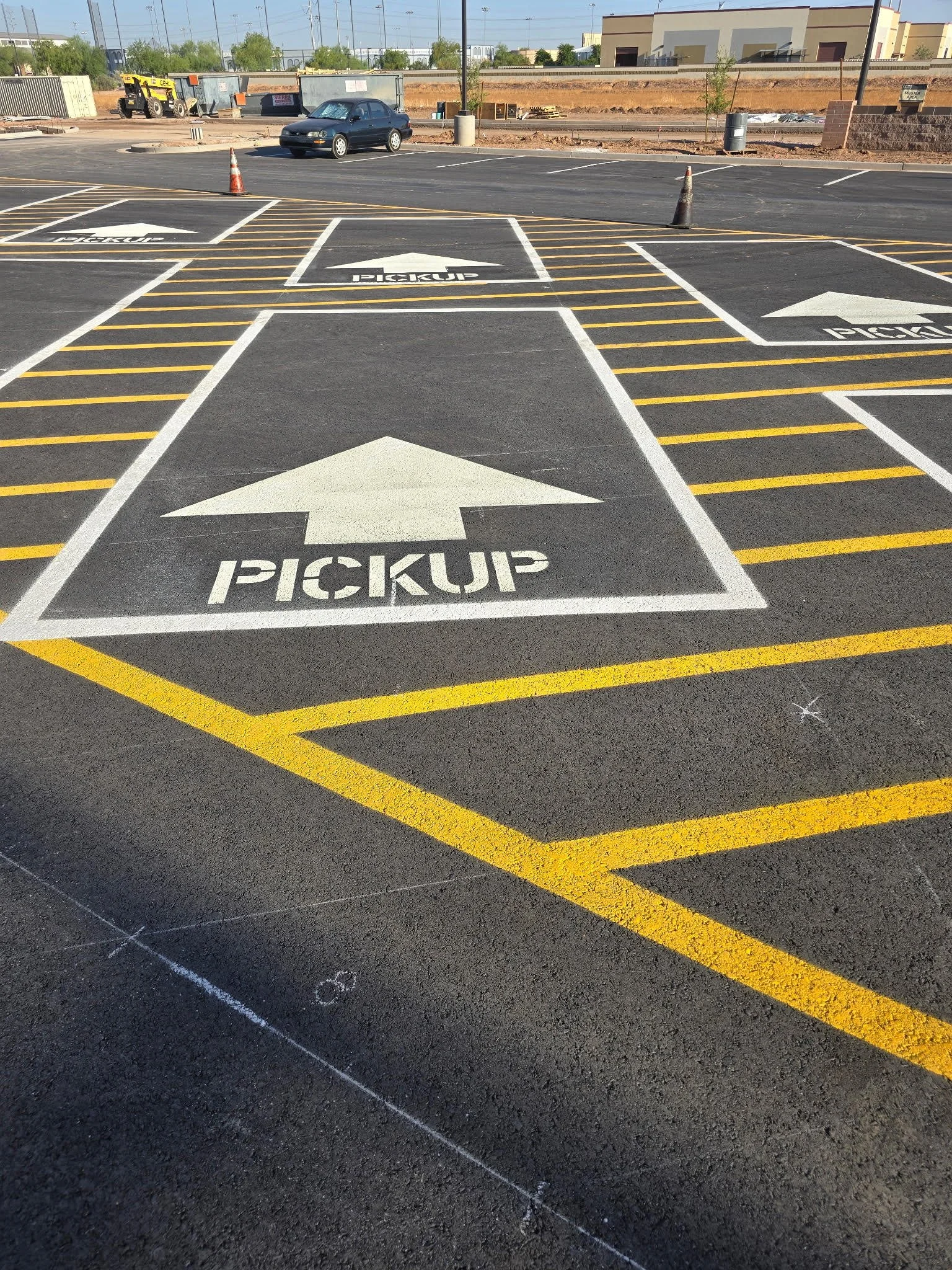 A parking lot with marked pickup zones, each with the word 'pickup' and an upward arrow, outlined in yellow and white lines. There is a car parked in the background and orange traffic cones placed near the pickup signs.