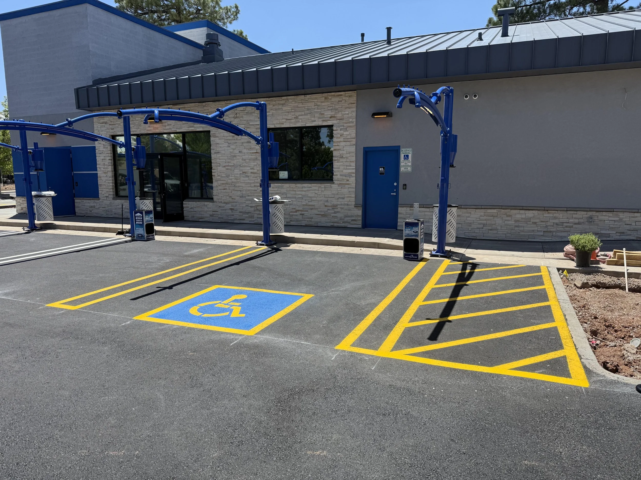 Parking lot with accessible parking space marked in blue and yellow, in front of a building with gray and brick exterior, blue doors, and blue overhangs, under clear sky and some trees.