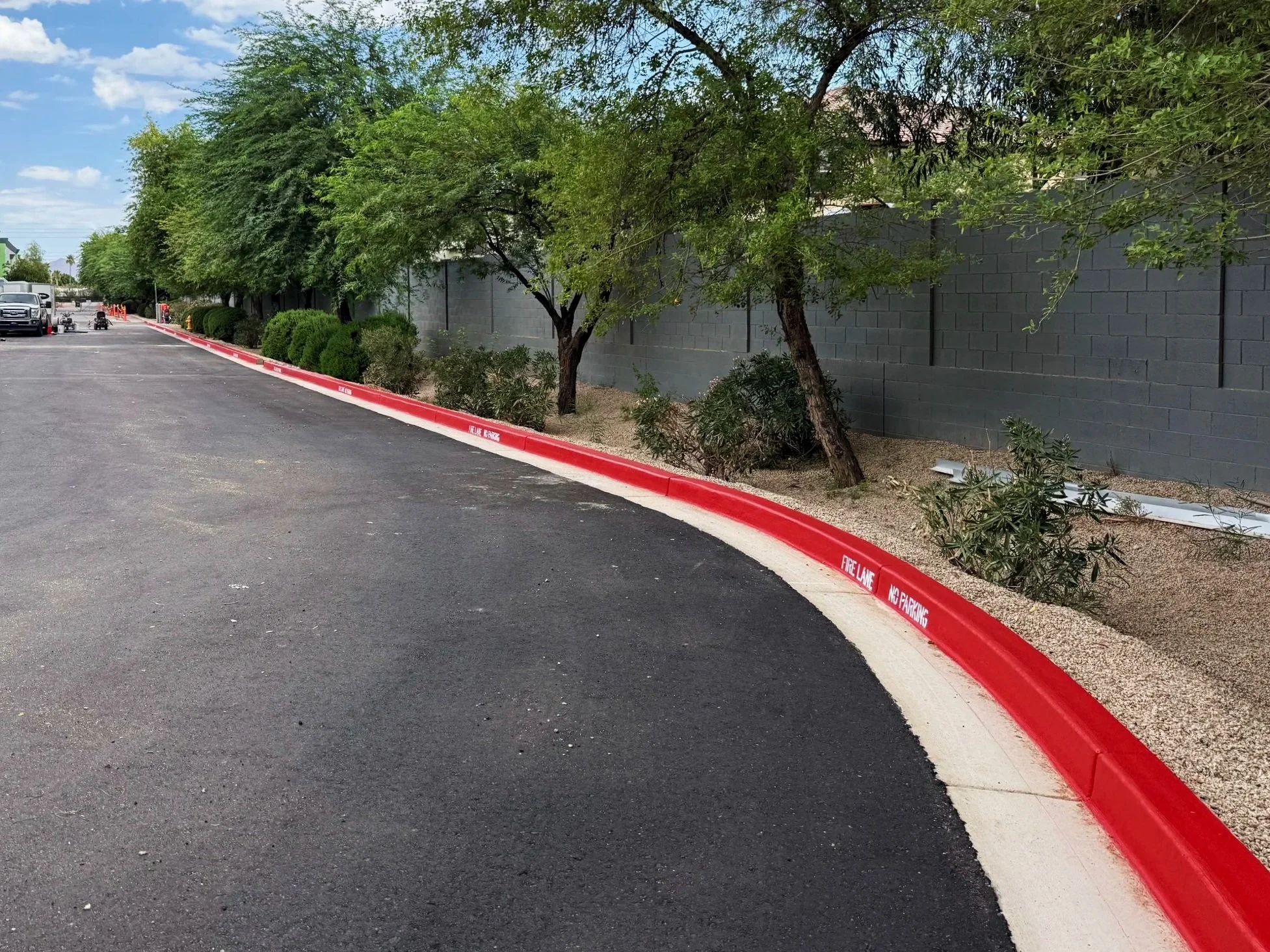 A parking lot with trees and bushes along a gray cinder block wall, and a red-painted curb with 'Fire Lane' and 'No Parking' written on it. Several parked vehicles are visible in the background.