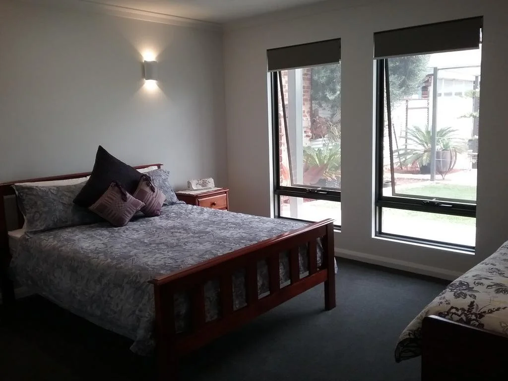 A bedroom with a large bed, pillows, a wooden nightstand, and two windows with a view of potted plants and a yard outside.