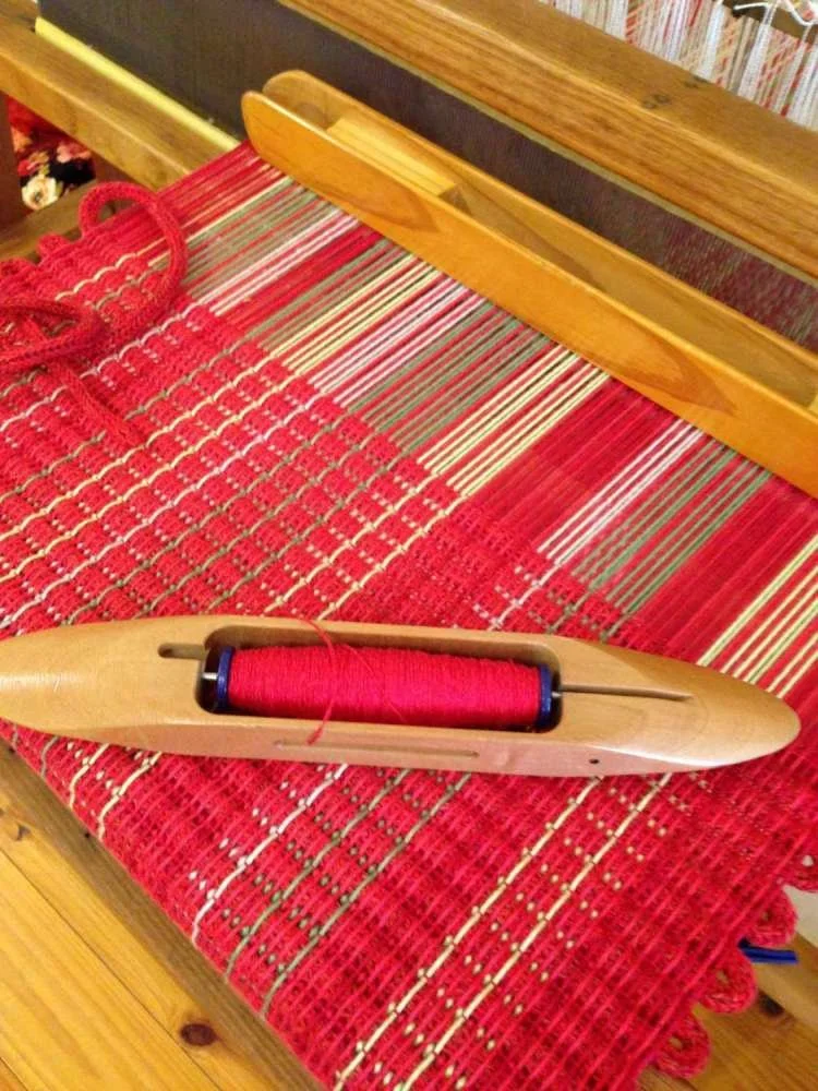 A vintage wooden weaving shuttle with red thread and a needle, resting on a red, green, and white woven textile on a wooden bench.