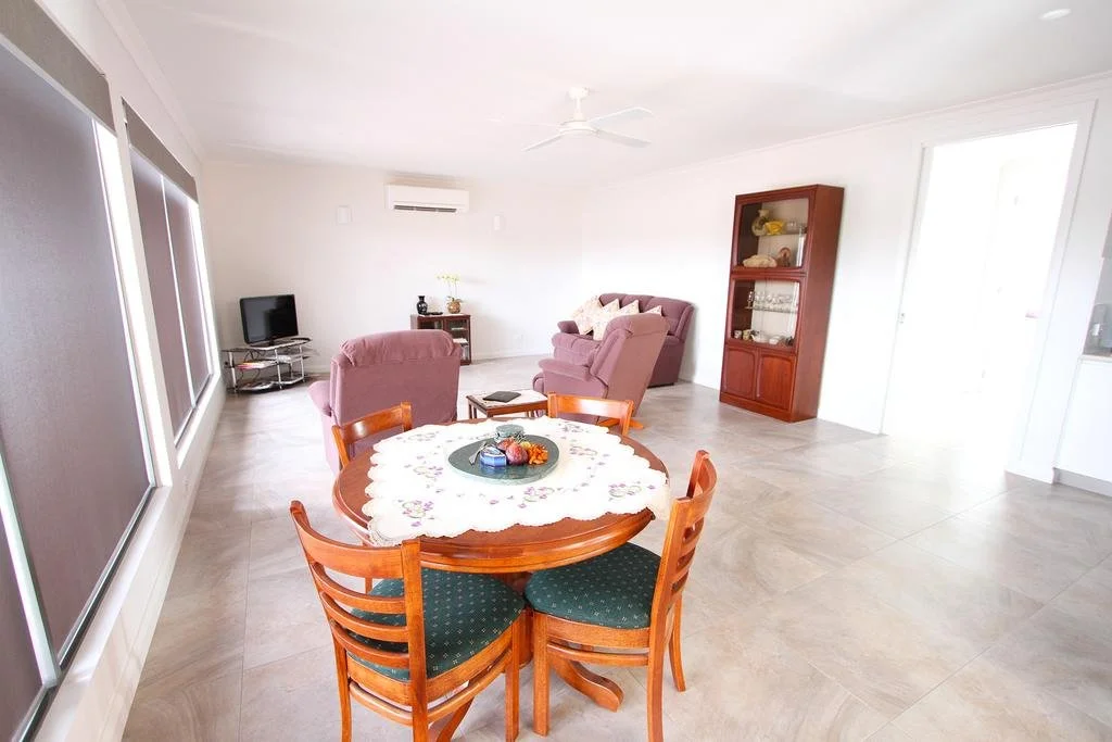 Spacious living room with a dining table, pink armchairs, a TV, and a display cabinet, with large windows and neutral-colored flooring.