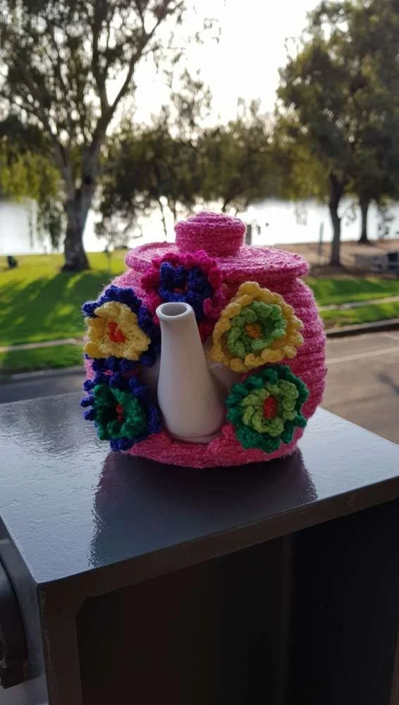 A teapot covered in bright pink yarn with colorful crocheted flowers, placed on a reflective surface outdoors with trees and water in the background.