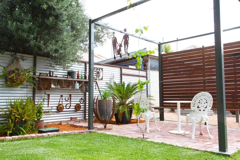 A backyard patio with a green lawn, potted plants, a white table, two ornate white chairs, decorative wall hanging, and a metal railing with silhouette figures of a family.