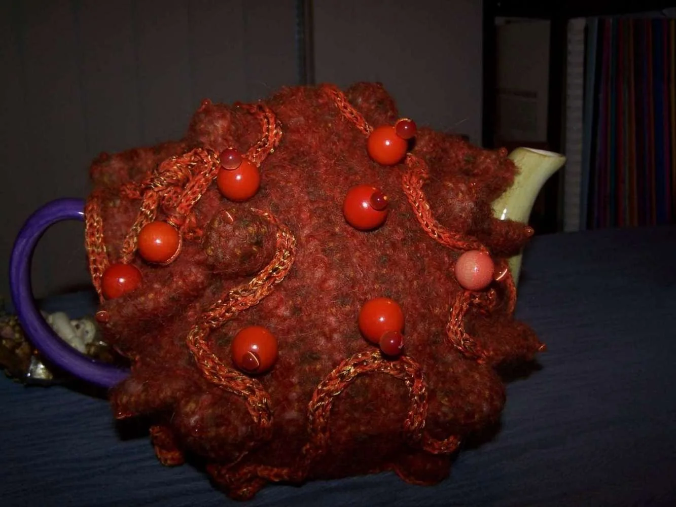 Decorative teapot with a fuzzy, reddish-brown surface, adorned with orange beads and braided orange string, placed on a dark table.