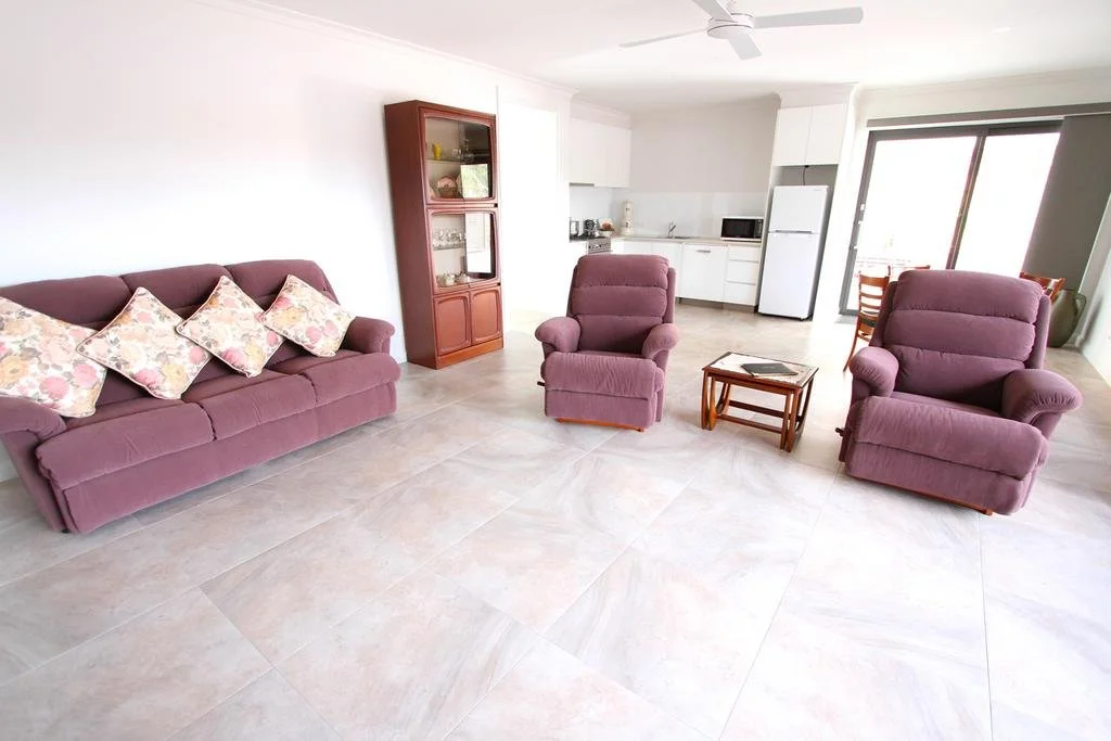 Living room with a purple sofa and two matching recliners, a glass-fronted wooden display cabinet, small wooden side table, and a kitchen area in the background. Large sliding glass door leading outside.