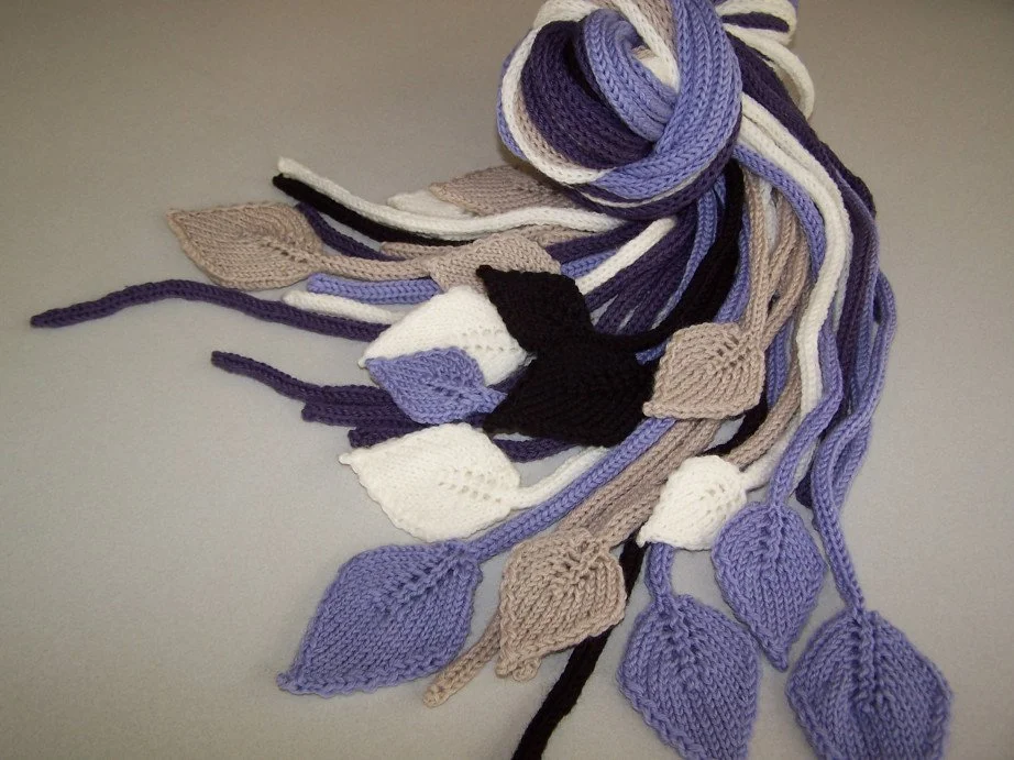 A bunch of knitted scarves in various colors including purple, white, black, gray, and beige, laid out on a flat surface.