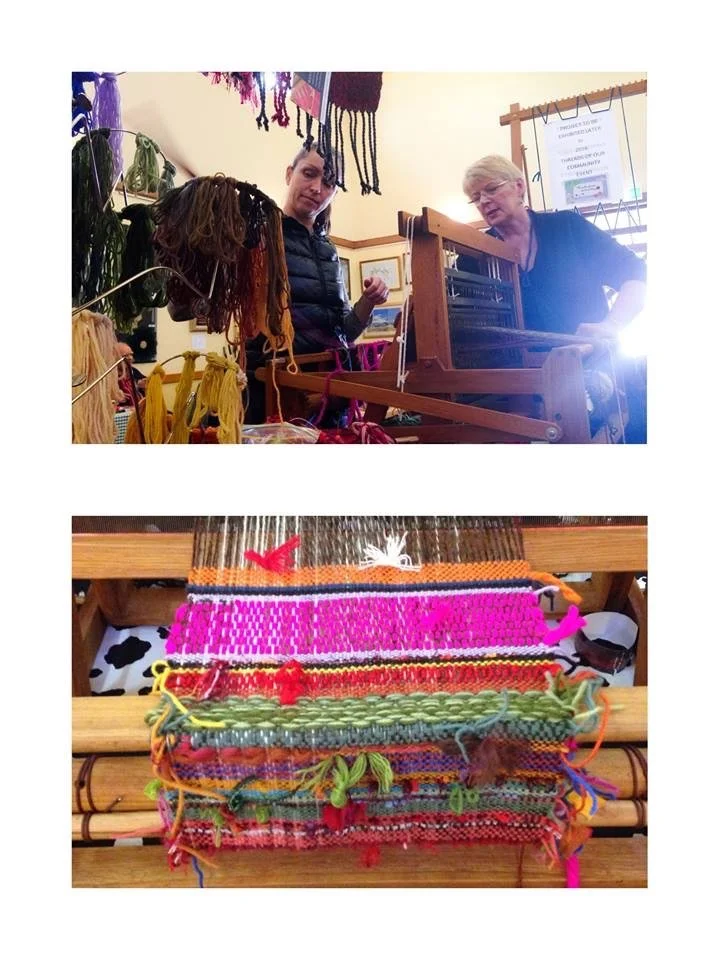 Two women at a craft fair, one using a spinning wheel and yarn fibers hanging on a rack, while the other observes; a colorful woven textile on a loom in the foreground.
