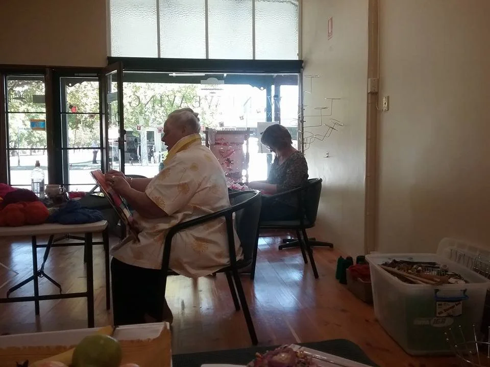 Two people are seated and reading inside a cafe or shop with large front windows, one man in a white Hawaiian shirt and one woman with short hair in a patterned top. The scene appears bright with natural light.