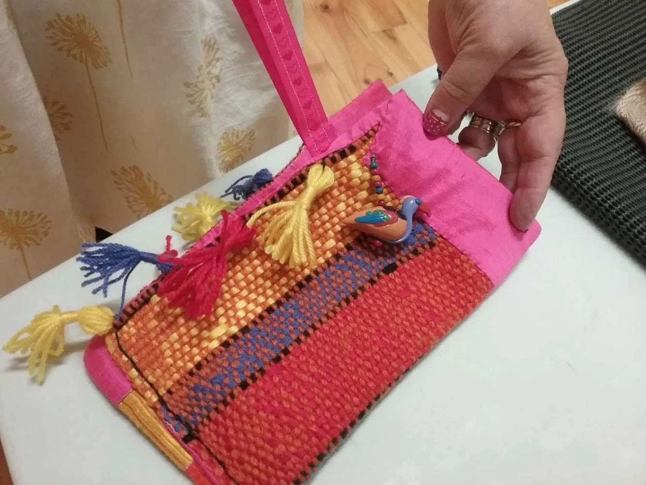 A person's hand holding a small, colorful woven bag decorated with tassels, a ceramic duck figurine, and a yellow piece of yarn. The bag has vibrant pink, orange, yellow, blue, and black colors and is resting on a white surface.
