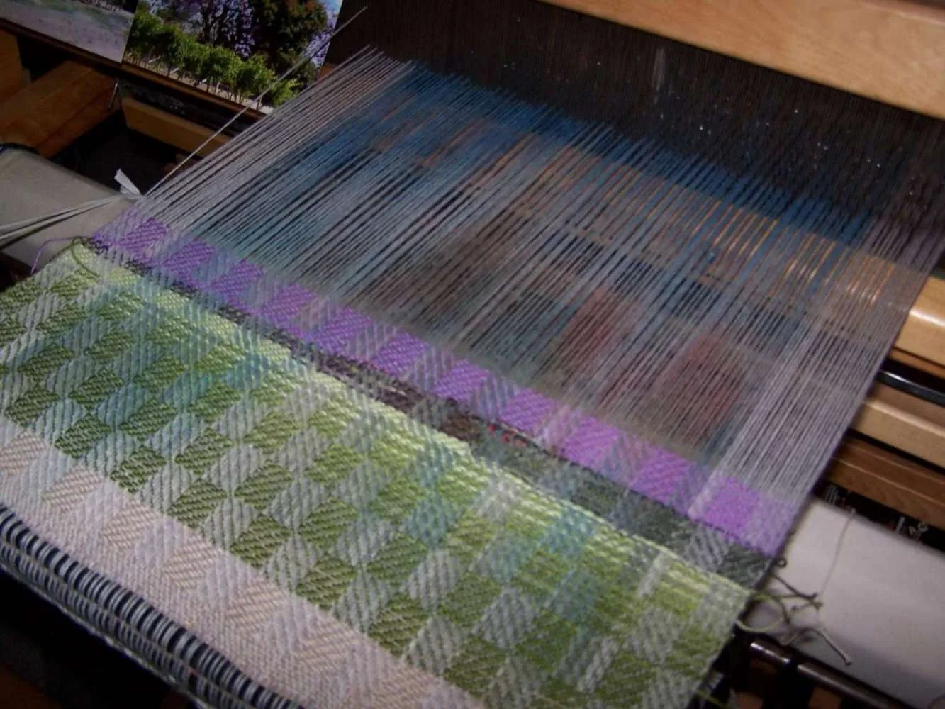 A loom with partially woven fabric featuring green and white diagonal checks and a section of vertical threads. A photo of a landscape with trees is partially visible in the background.