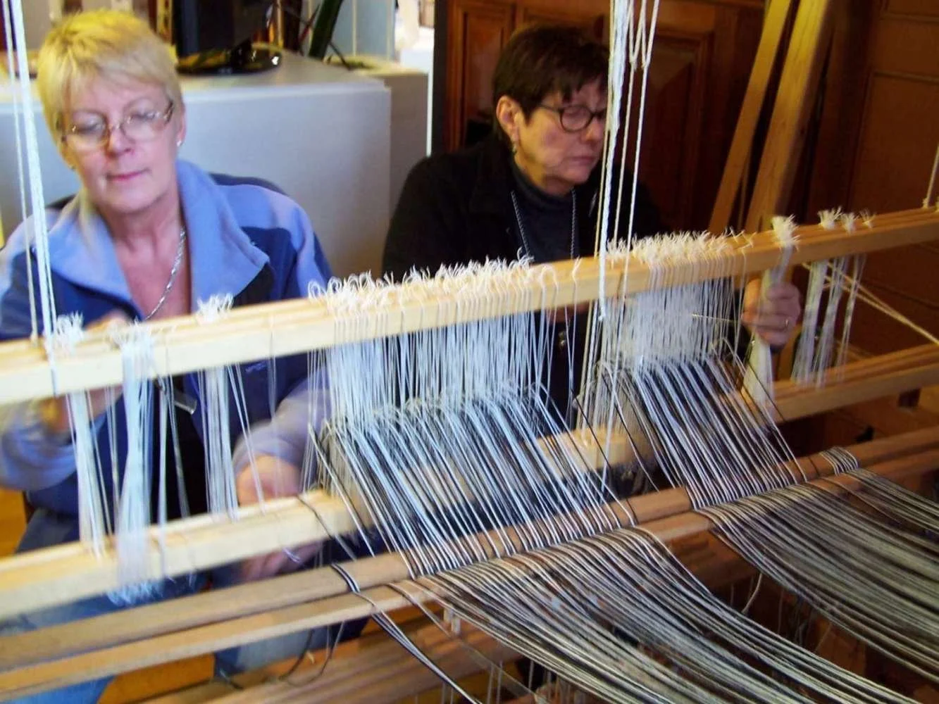 Two women working on a traditional loom weaving fabric, with wooden parts and numerous threads.
