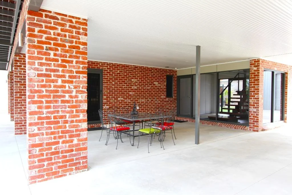 Covered outdoor patio with red brick walls, a black metal dining table, six chairs with red and green cushions, and a staircase visible through glass doors.