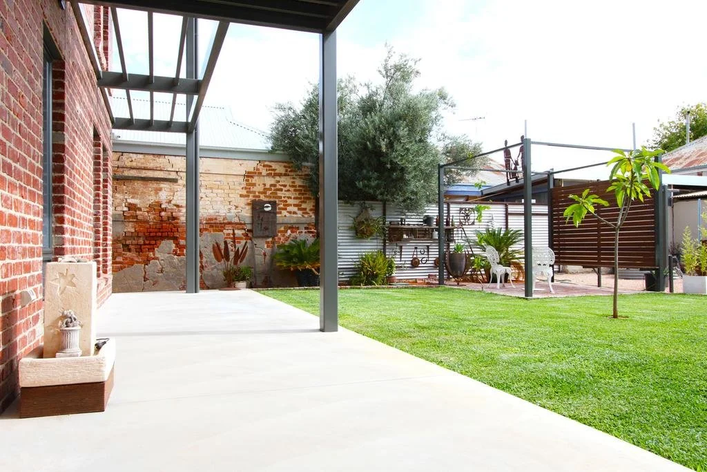 A backyard patio with a concrete floor, a brick wall on the left, and a garden with green grass, trees, and potted plants. There are outdoor chairs and shade structures.