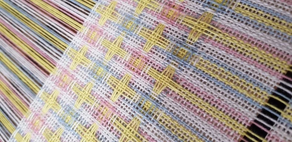 Close-up of colorful woven fabric with pink, yellow, and blue threads and intricate cross-stitch pattern.