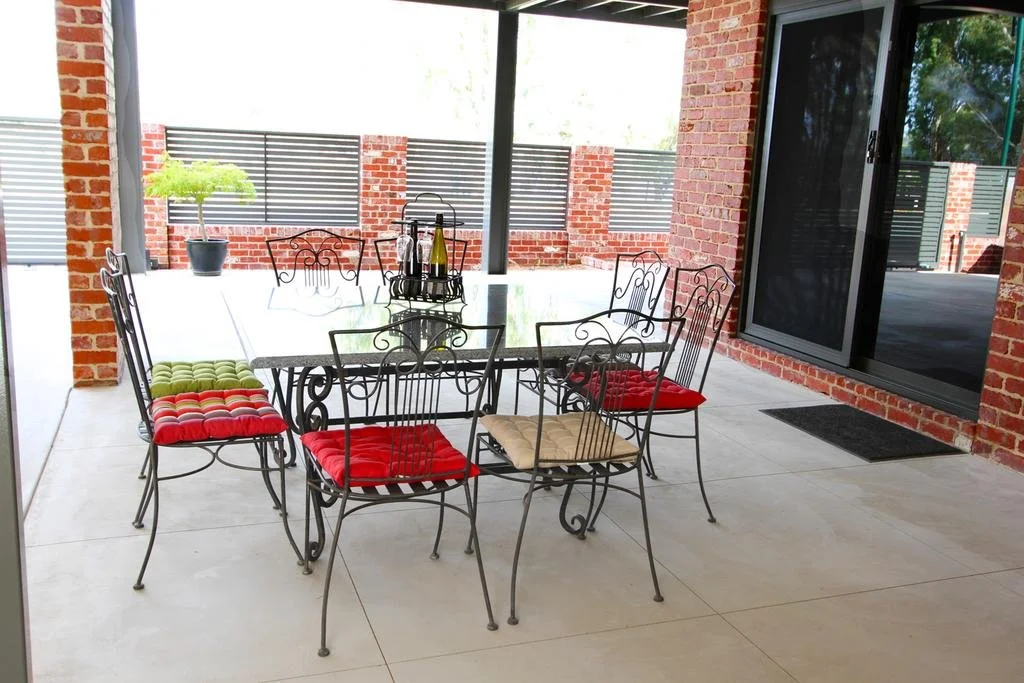Outdoor patio area with a black metal dining table, six matching chairs with red, green, and beige cushions, a potted plant, and decorative bottles on a tray. There's brick walls, tiled flooring, and sliding glass doors.