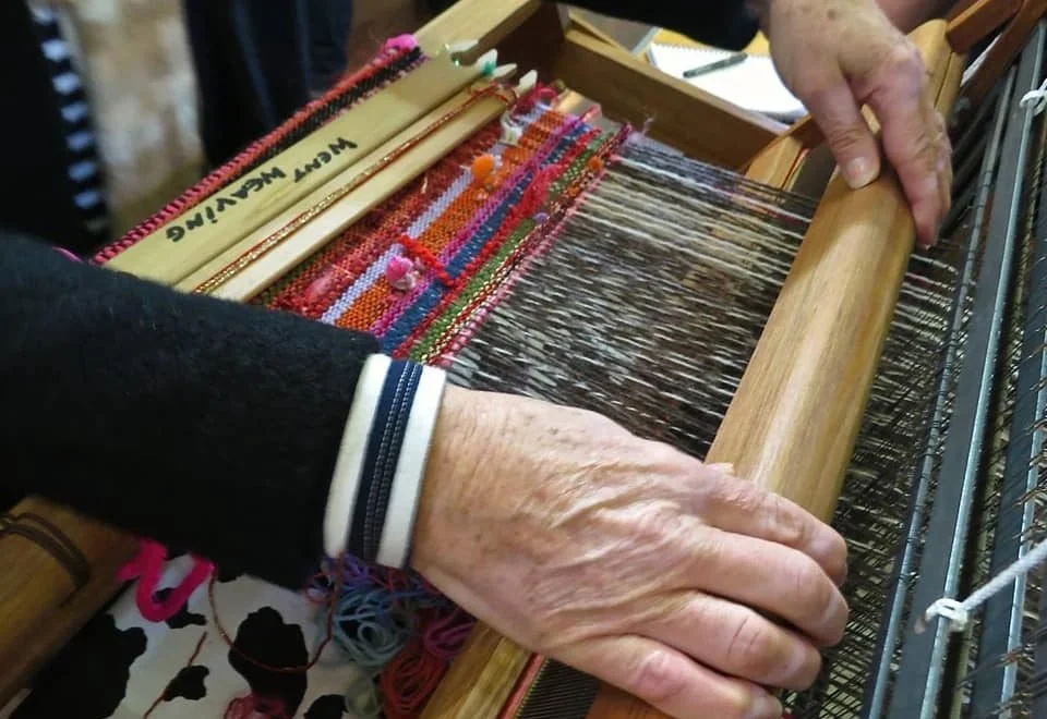 Close-up of elderly hands operating a weaving loom with colorful threads, producing a vibrant woven fabric.