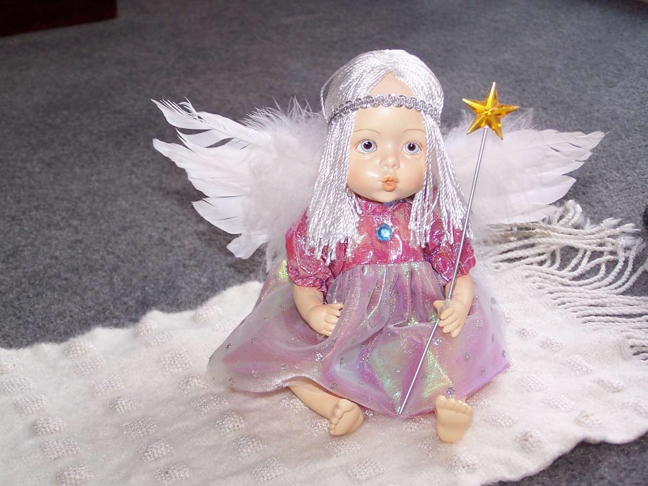 A doll with angel wings, white hair, and a silver crown, wearing a pink dress with a glittery iridescent overlay, holding a small magic wand with a gold star, sitting on a white textured surface.