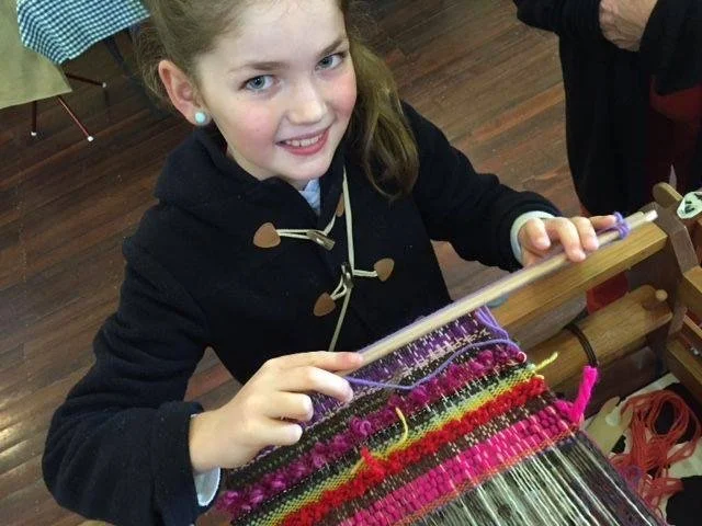 A young girl with light brown hair working on a colorful weaving loom, smiling at the camera.