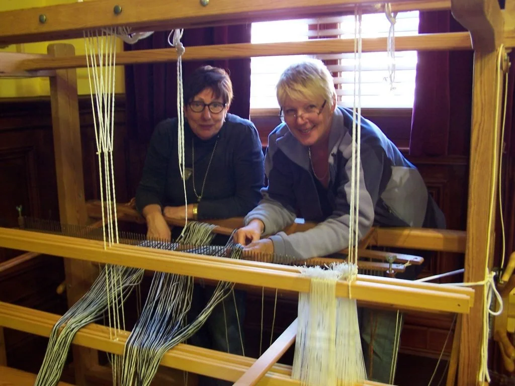 Two women using a large wooden loom to weave fabric in a room with wooden walls and a window.