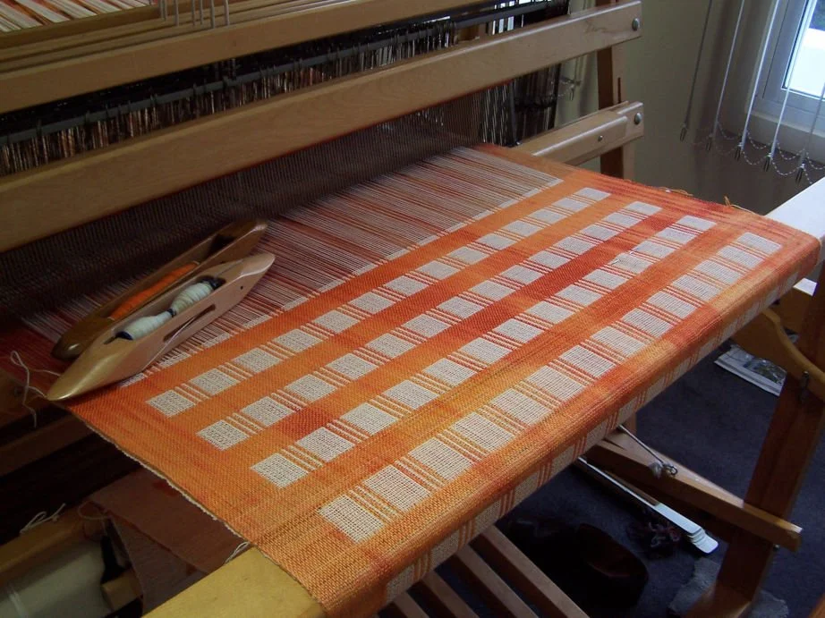A weaving loom with an orange and beige checked woven fabric in progress, a shuttle, and white thread spools on a table near a window.