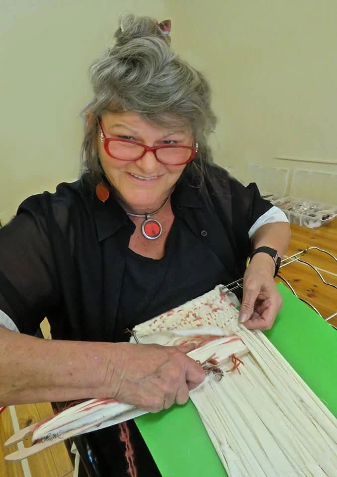 Older woman with gray hair, red glasses, and colorful earrings, weaving or crafting with fabric and yarn on a green table, smiling at the camera.
