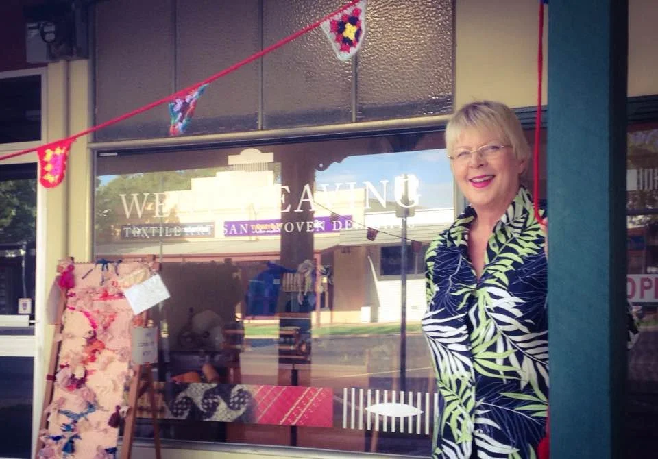 A smiling woman with short blond hair, glasses, wearing a dark shirt with green and white leaf patterns, standing outside a store with a glass window. The store has decorations including a display of fabric with floral designs and colorful bunting ha