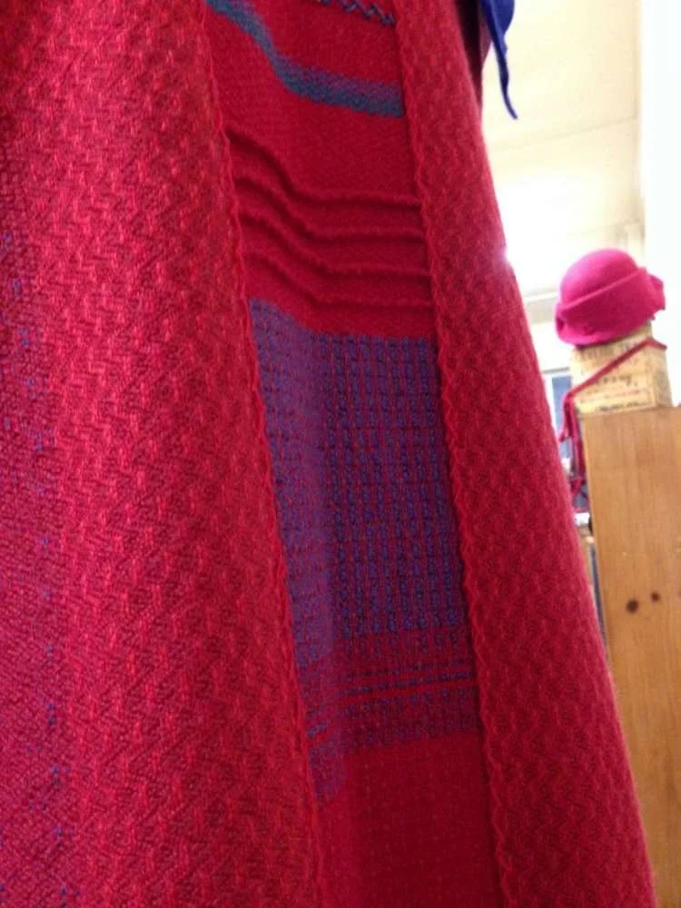 A person is wearing a red textured coat and a red and purple patterned sweater. In the background, there is a pink hat on a mannequin head and some wooden furniture.