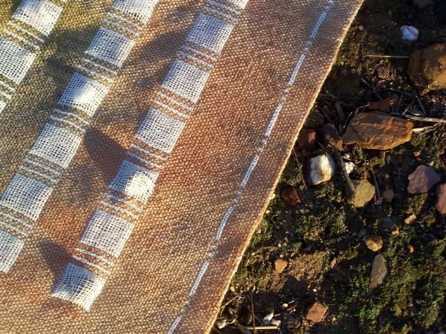 Close-up of a woven fabric with brown, white, and blue patterns, placed on the ground with rocks and small plants.