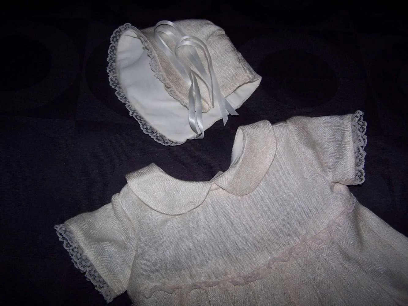 A white dress with lace trim and a matching bonnet with lace trim, displayed on a dark surface.