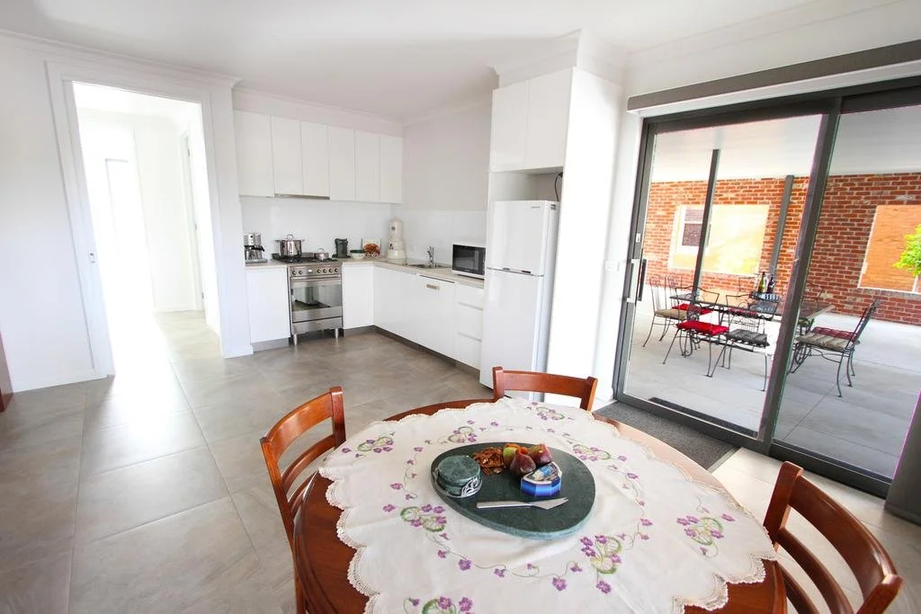 Kitchen and dining area with a round table, four wooden chairs, a white countertop, white cabinets, a sliding glass door leading to a patio with outdoor furniture, and brick wall in the background.