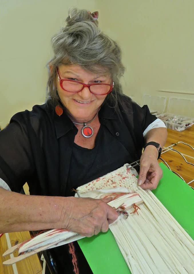 Older woman with gray hair, red glasses, and a black shirt, smiling while weaving fabric on a loom.