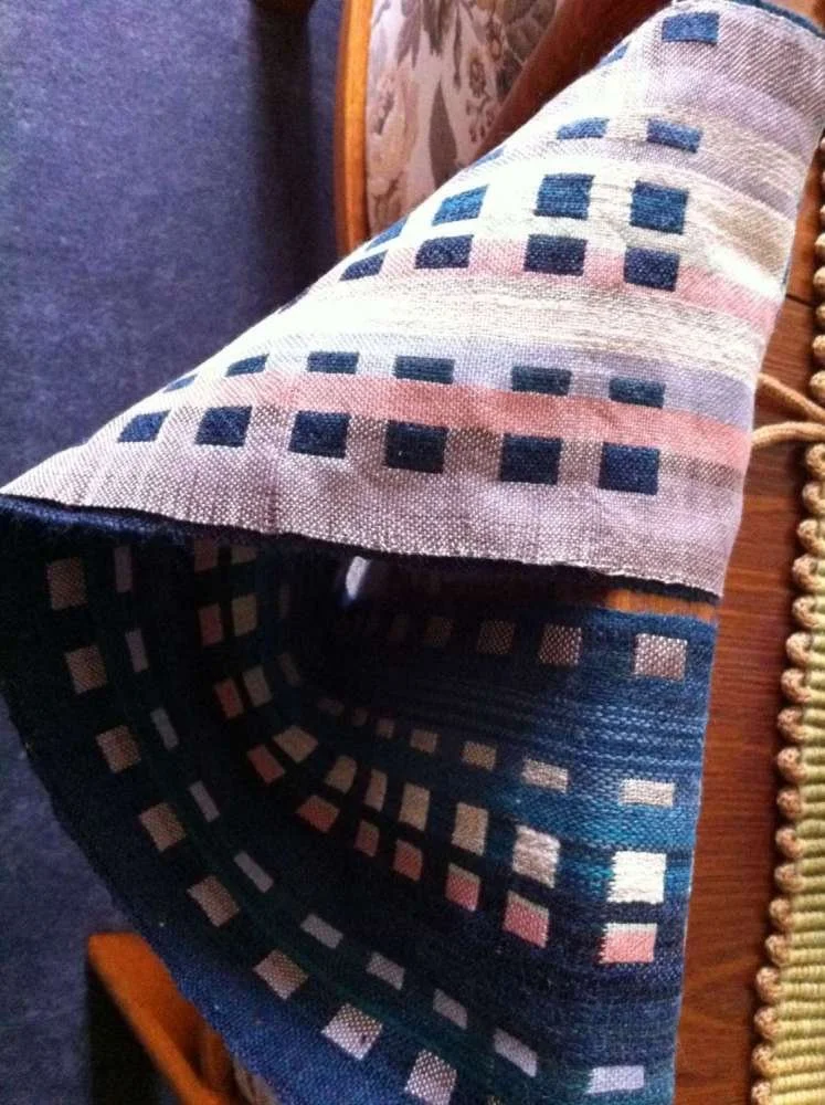 Close-up of a woven textile with geometric square pattern in shades of pink, blue, cream, and black, partially draped over a piece of furniture.