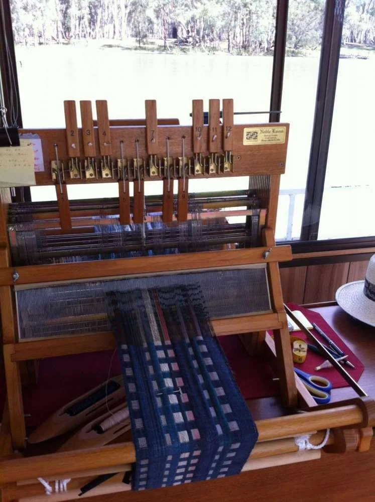 Loom for weaving with partially woven blue fabric, tools and supplies nearby, large window with a view of trees outside.