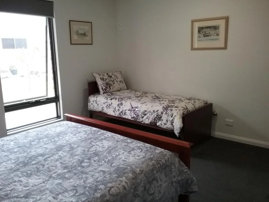 A bedroom with two beds, one with floral bedding near a window and the other with a patterned coverlet, with framed pictures on the wall.