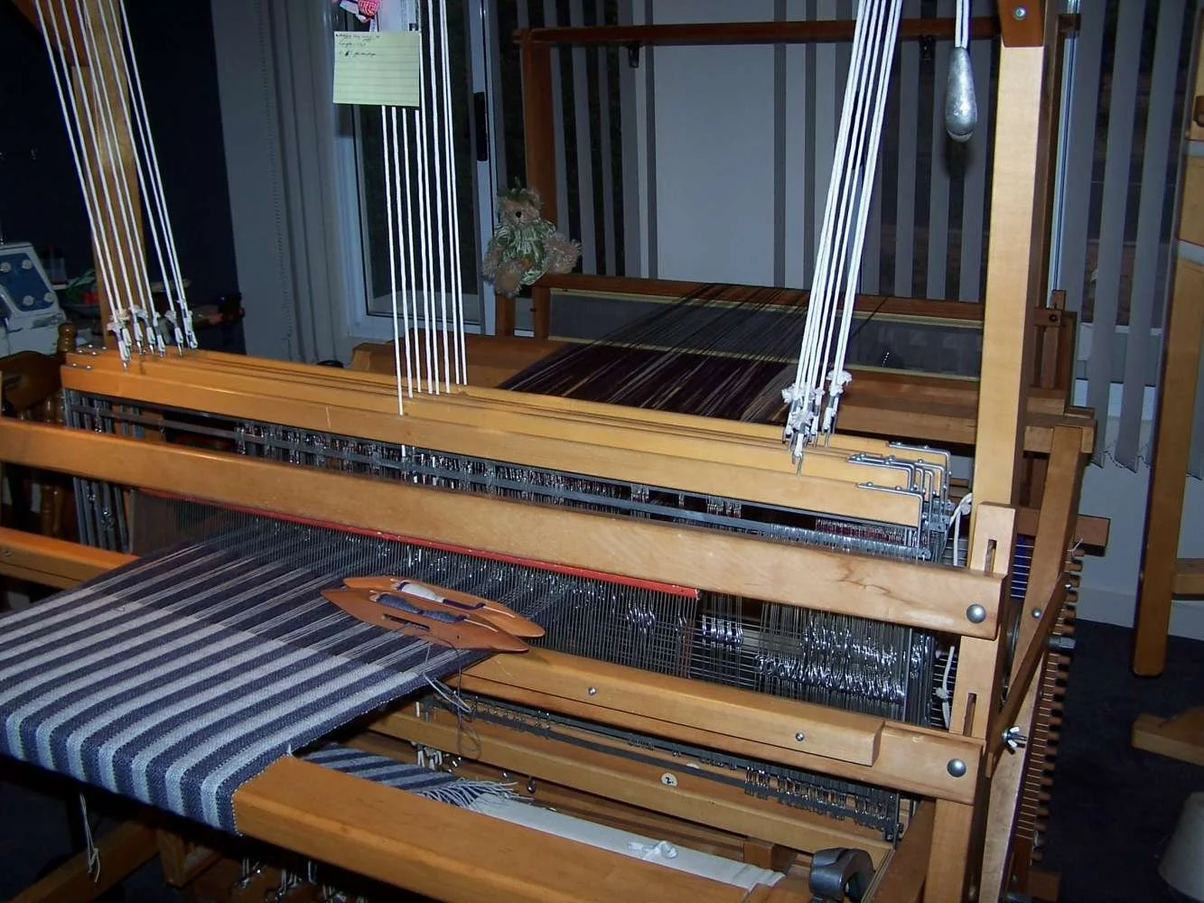 A large, wooden loom with multiple parts and fabric woven on it, situated in a room with a window in the background.