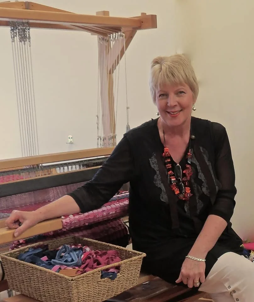 A woman with short blonde hair sitting next to a large weaving loom, with a basket of colorful yarns in front of her.