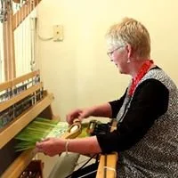A woman sitting at a loom or weaving machine, working with green yarn in a well-lit room.