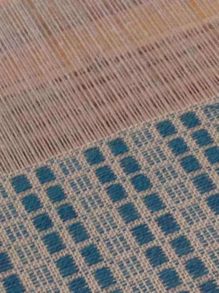 Close-up of a woven textile with a pattern of blue squares and beige grid lines next to a wooden surface with vertical grain.