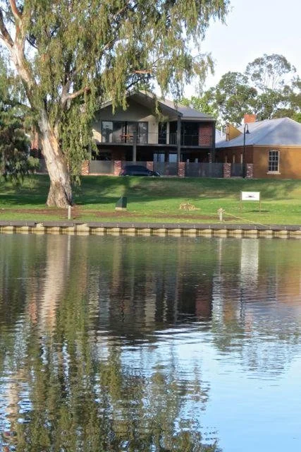 Modern house with large balcony, trees, and a boat dock along a river in a suburban neighborhood.