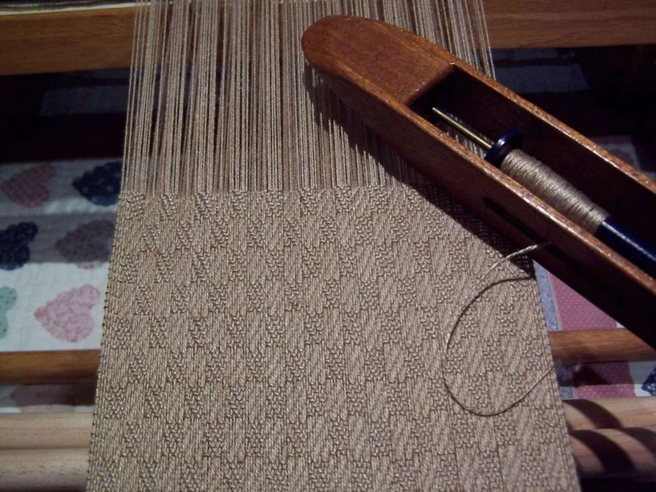 A weaving loom with a partially woven fabric in beige and light brown colors. The loom holds a spool of thread and the woven fabric is textured with a pattern of small, tightly woven stitches.