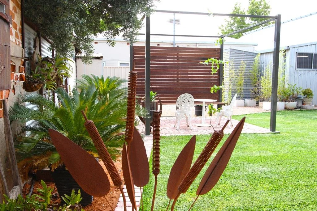 A backyard garden with lush green grass, potted plants, and outdoor furniture, including white chairs and a round table, all under a black metal frame.