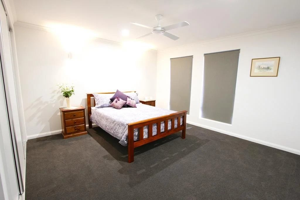A bedroom with a wooden bed frame, gray wallpaper, gray carpeting, two nightstands, a ceiling fan, and two windows with gray blinds.