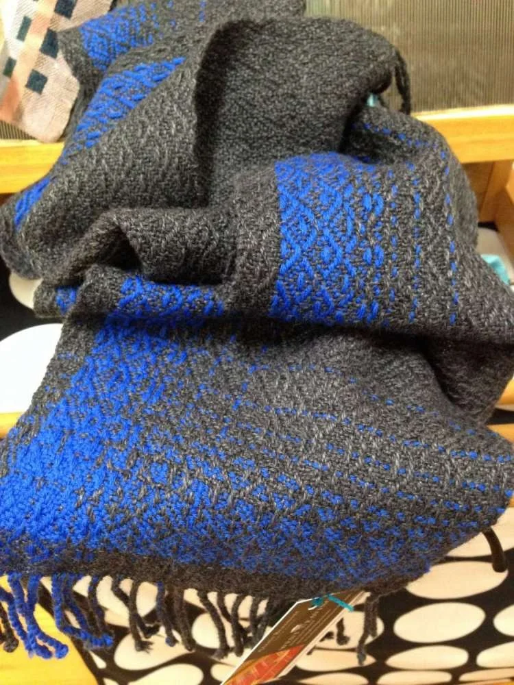 Close-up of a folded black and blue textured knit scarf with fringe at the ends, resting on a surface with a black and white polka dot pattern.