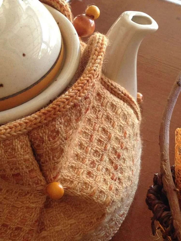 Close-up of a white teapot with a spout, wrapped in a beige crocheted cover with yellow beads.