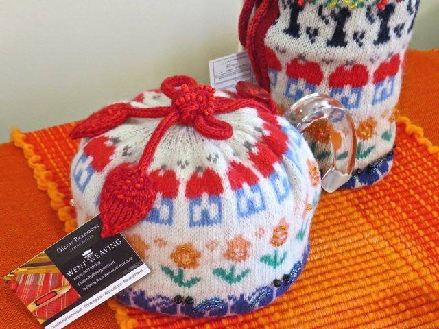 Knitted hot water bottle covers with colorful patterns, one with a red drawstring and orange heart, placed on an orange textured surface.