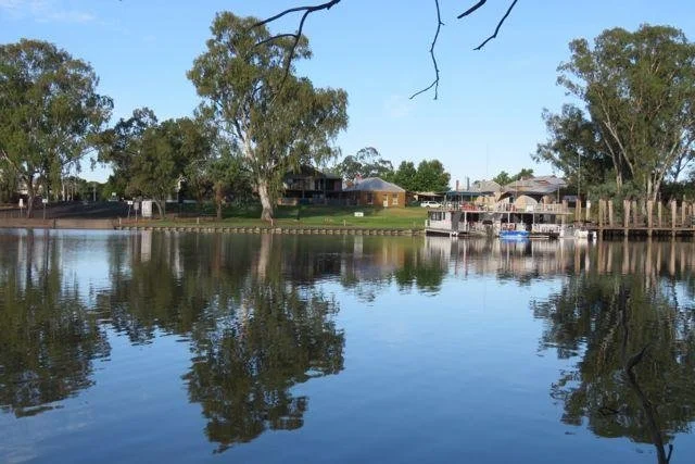 Calm river or lake with reflections of trees and houses on the opposite bank, clear blue sky overhead.