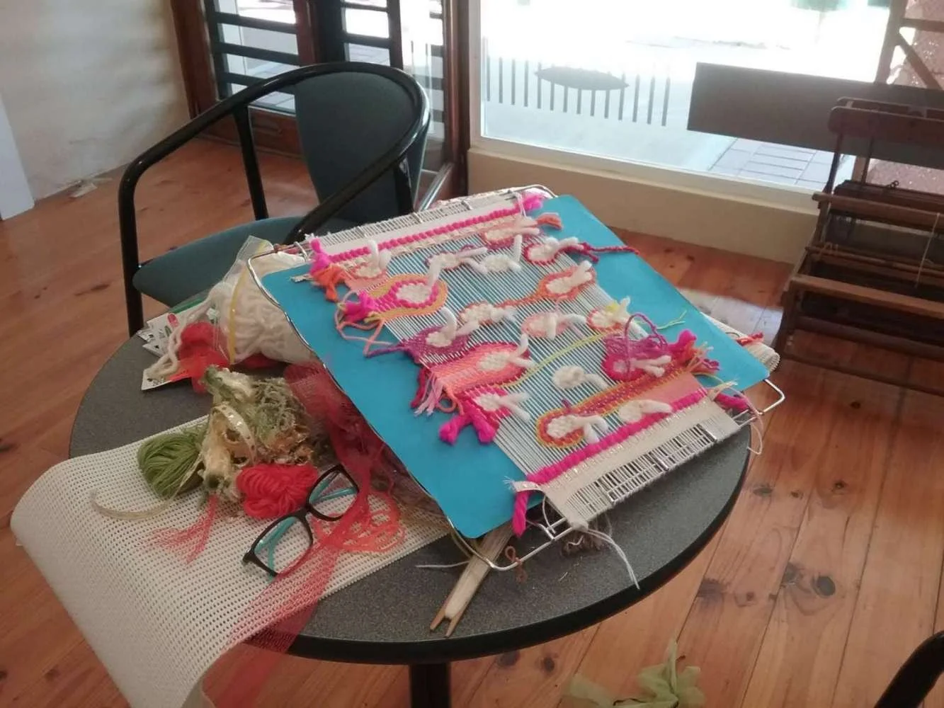 A table with colorful woven wall hangings, with various yarns, a pair of glasses, and crafting supplies in a room with wooden floors and large windows.